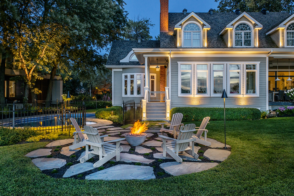 Vue nocturne d'un espace extérieur avec chaises autour d'un feu, maison en arrière-plan